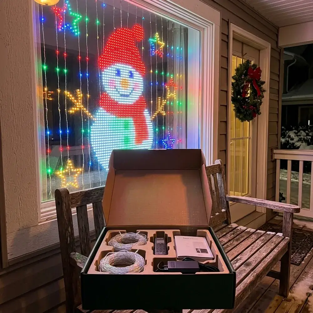 Caja del producto abierta en el porche y la ventana con un muñeco de nieve dibujado por las luces